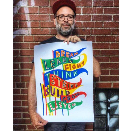 Artist and designer Josh MacPhee holding a poster depicting pennants that list the words dream, learn, fight, think, strike, build, grow, listen